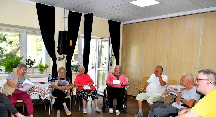 Grupa seniorów siedzi w kręgu na sali podczas zajęć. Uczestnicy trzymają w rękach kartki i rozmawiają, uśmiechając się. W tle widoczne są stoły, kwiaty na parapecie oraz otwarte drzwi.