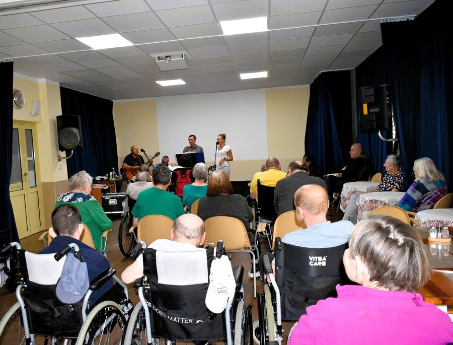 Grupa mieszkańców, część z nich na wózkach inwalidzkich, siedzi w sali i ogląda występ muzyczny. Na scenie stoi troje wykonawców: dwóch gitarzystów i wokalistka z mikrofonem.