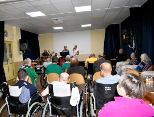 Grupa mieszkańców, część z nich na wózkach inwalidzkich, siedzi w sali i ogląda występ muzyczny. Na scenie stoi troje wykonawców: dwóch gitarzystów i wokalistka z mikrofonem.