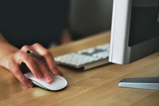 Zdjęcie przedstawia zbliżenie dłoni osoby trzymającej mysz komputerową na drewnianym biurku. Osoba ta trzyma również długopis między palcami. W tle znajduje się klawiatura oraz monitor.