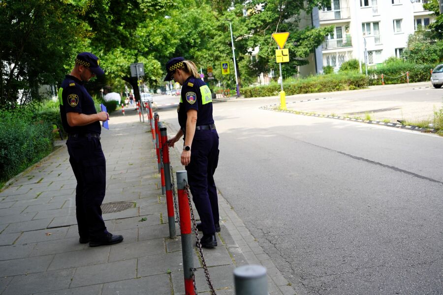 Dwóch strażników miejskich w mundurach stoi na chodniku przy ulicy. Jeden trzyma notatnik, drugi ogląda słupek z łańcuchem. W tle widać drzewa, znaki drogowe i budynki mieszkalne. Scena odbywa się w s