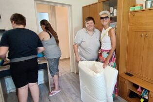 Uśmiechnięte kobiety z materacem na tle mebli oraz stojącej tyłem matki oraz syna.