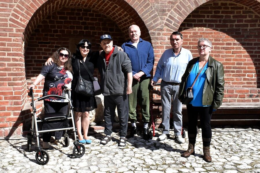 Na zdjęciu widzimy sześć osób stojących na tle czerwonej ceglanej ściany z łukowymi wnękami. Grupa pozuje do wspólnej fotografii w słoneczny dzień. 