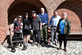Barbakan 26.08.2025 Na zdjęciu widzimy sześć osób stojących na tle czerwonej ceglanej ściany z łukowymi wnękami. Grupa pozuje do wspólnej fotografii w słoneczny dzień.