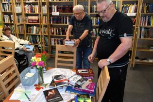 Biblioterapia 26.08.2025 Na zdjęciu widzimy wnętrze biblioteki. Przy stole z obrusem i wazonem kwiatów leży kilka książek, m.in. biografie i powieści. Dwaj starsi mężczyźni stoją obok stołu.