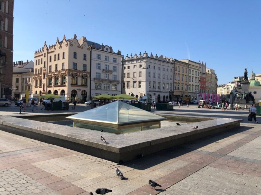 Zdjęcie przedstawia fontannę na Rynku Głównym w Krakowie. Oszklona fontanna w kształcie piramidy, w tle kamienice oraz pomnik Adama Mickiewicza.