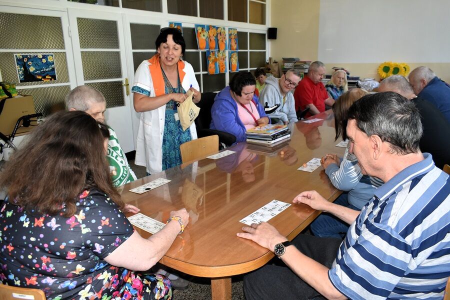 Grupa osób siedzi przy długim stole, biorąc udział w zajęciach – grają w lotto, a prowadząca w białym fartuchu z pomarańczowym kołnierzem trzyma woreczek z baryłkami.