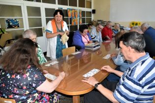 Turniej lotto 27.08.2025 Grupa osób siedzi przy długim stole, biorąc udział w zajęciach – grają w lotto, a prowadząca w białym fartuchu z pomarańczowym kołnierzem trzyma woreczek z baryłkami.
