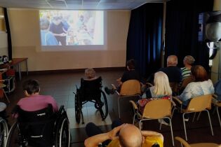 Zajęcia kronikarskie 28.08.2025 Grupa osób siedzi na krzesłach i w wózkach inwalidzkich w sali, oglądając projekcję zdjęć i filmów z wycieczek na dużym ekranie.