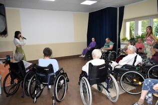 Grupa osób starszych, głównie na wózkach inwalidzkich, siedzi w sali i ogląda występ kobiety śpiewającej do mikrofonu.