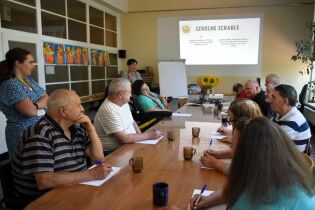 Seniorzy uczestniczą w zajęciach poznawczych przy stole, zapisując odpowiedzi w zeszytach. Na ekranie wyświetlony jest slajd z napisem „Szkolne Scrabble”.