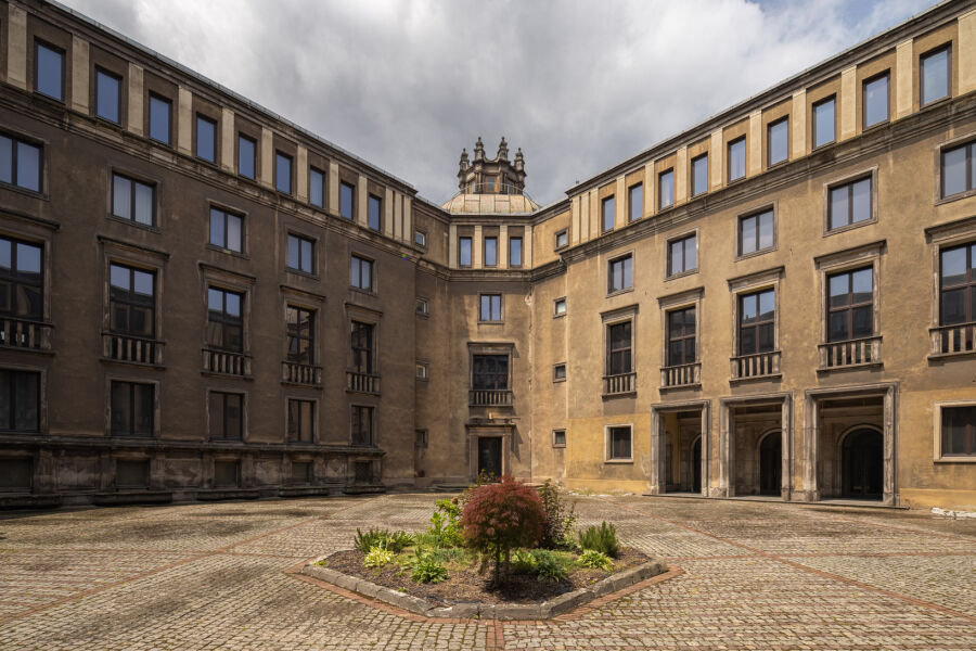Dziedziniec sfotografowany w lekko pochmurny dzień. Na pierwszym planie otwarta przestrzeń oraz dwie ściany budynku z czerwonej cegły, łączące się w narożniku. Podkreślona faktura muru i architektura.