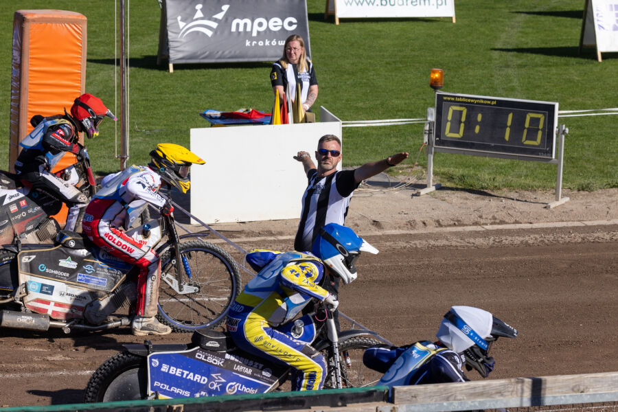 Na torze żużlowym zawodnicy na motocyklach szykują się do startu. Arbiter w czarno-białej koszulce daje sygnał ręką. W tle widoczna jest elektroniczna tablica z czasem oraz zielona murawa.