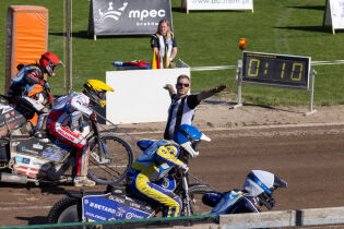 Na torze żużlowym zawodnicy na motocyklach szykują się do startu. Arbiter w czarno-białej koszulce daje sygnał ręką. W tle widoczna jest elektroniczna tablica z czasem oraz zielona murawa.