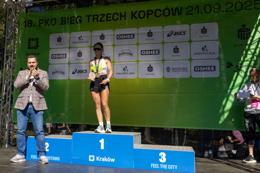 Na zdjęciu widać podium z biegaczką która zajęła 1 miejsce. W tle widać baner z logami sponsorów i nazwą biegu - 18. PKO Bieg Trzech Kopców.