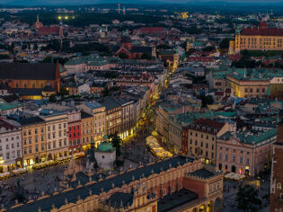 Fotografia centrum Krakowa z drona wykonana wieczorem. Widoczne stare miasto z oświetlonym rynkiem w centrum oraz promienisty układ ulic ścisłego centrum. 