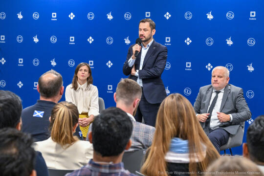 Fotografia ze spotkania Prezydenta Krakowa z cudzoziemcami. W centrum zdjęcia Prezydent Aleksander Miszalski przemawia stojąc i trzymając mikrofon bezprzewodowy. W pierwszym planie głowy uczestników. 