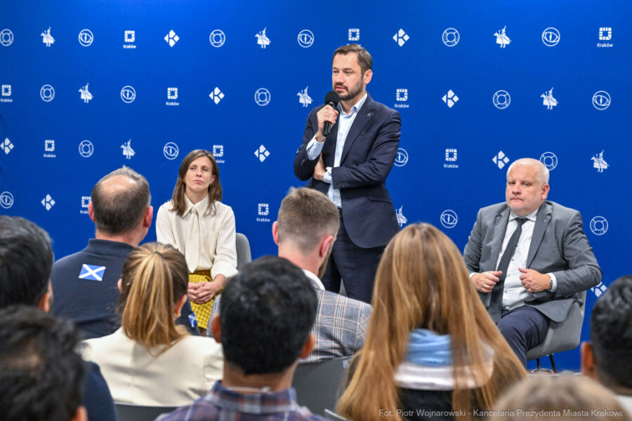 Fotografia ze spotkania Prezydenta Krakowa z cudzoziemcami. W centrum zdjęcia Prezydent Aleksander Miszalski przemawia stojąc i trzymając mikrofon bezprzewodowy. W pierwszym planie głowy uczestników. 