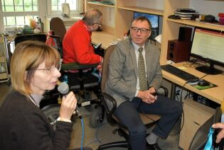 Audycja radiowęzła 26.09.2025 Mężczyzna w garniturze i kobieta z mikrofonem siedzą przy komputerach w pokoju radiowęzła, obok nich mężczyzna na wózku inwalidzkim.