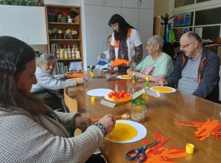 Arteterapia 26.09.2025 Grupa seniorów siedzi przy stole i wykonuje prace plastyczne z kolorowego papieru pod opieką instruktorki arteterapii.