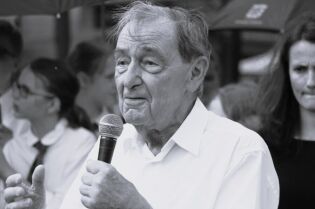 czarno-białe zdjęcie przedstawia zbliżenie Pastora Friedricha Magiriusa przemawiającego na wydarzeniu plenerowym. W lewej ręce trzyma mikrofon bezprzewodowy. W tle uczestnicy spotkania.   