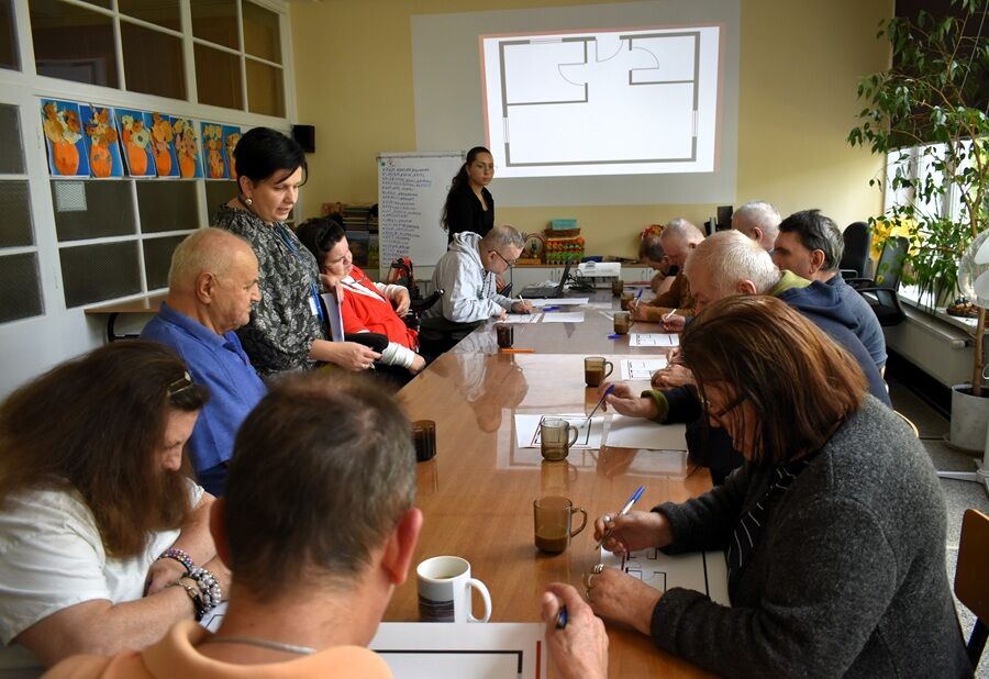 Grupa osób siedzi przy długim stole w sali, uczestnicząc w zajęciach edukacyjnych. Prowadząca stoi przy tablicy z rzutem prostego planu pomieszczenia. Uczestnicy wypełniają karty pracy i rozmawiają. 