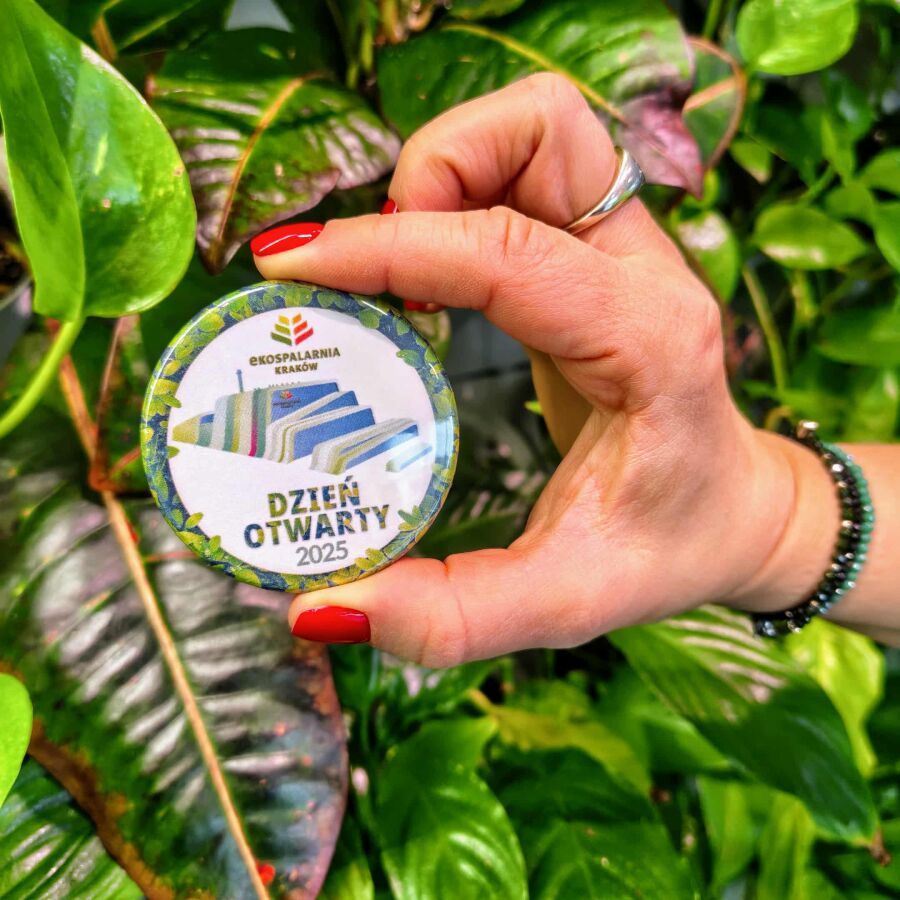 Zdjęcie przedstawia rękę kobiety trzymającej w palcach odznakę z dnia otwartego w Ekospalarni. W tle widać zieloną roślinę.
