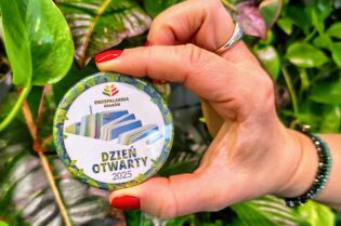 Zdjęcie przedstawia rękę kobiety trzymającej w palcach odznakę z dnia otwartego w Ekospalarni. W tle widać zieloną roślinę.