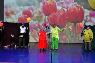 Spotkania Artystyczne 22.10.2025 Grupa aktorów występuje na scenie w kolorowych kostiumach przedstawiających owady i kwiaty. W tle widać duży ekran z obrazem tulipanów.