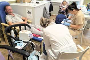 Mężczyzna siedzi w fotelu i uśmiecha się, podczas gdy dwie pielęgniarki lub podolodzy zajmują się jego stopami. W tle widać inne osoby na wózkach inwalidzkich. Na stole obok stoją narzędzia medyczne. 