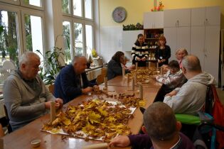 Grupa osób starszych siedzi przy dużym stole w jasnej sali i uczestniczy w zajęciach plastycznych. Na stole leżą jesienne liście i tekturowe rolki. Dwie instruktorki stoją i pomagają uczestnikom.