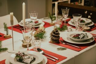 Grafika przedstawia świątecznie nakryty stół, z białym obrusem i czerwonymi serwetkami, na których stoją białe talerze, sztućce oraz szklanki i dekoracje.