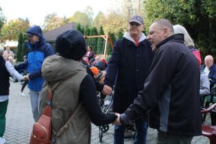 Grupa osób trzyma się za ręce w kręgu podczas zabawy na zewnątrz, w tle widać innych uczestników siedzących na ławkach.