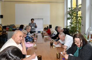 Grupa seniorów siedzi przy dużym stole w jasnej sali i uczestniczy w zajęciach prowadzonych przez kobietę stojącą przy tablicy. Uczestnicy piszą na kartkach, a na stole stoją kubki z napojami.