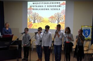 Grupa dzieci śpiewa do mikrofonów podczas występu na scenie. Za nimi widoczny jest ekran z napisem „Międzypokoleniowe spotkania z seniorami – Odblaskowa szkoła” oraz jesienna grafika.