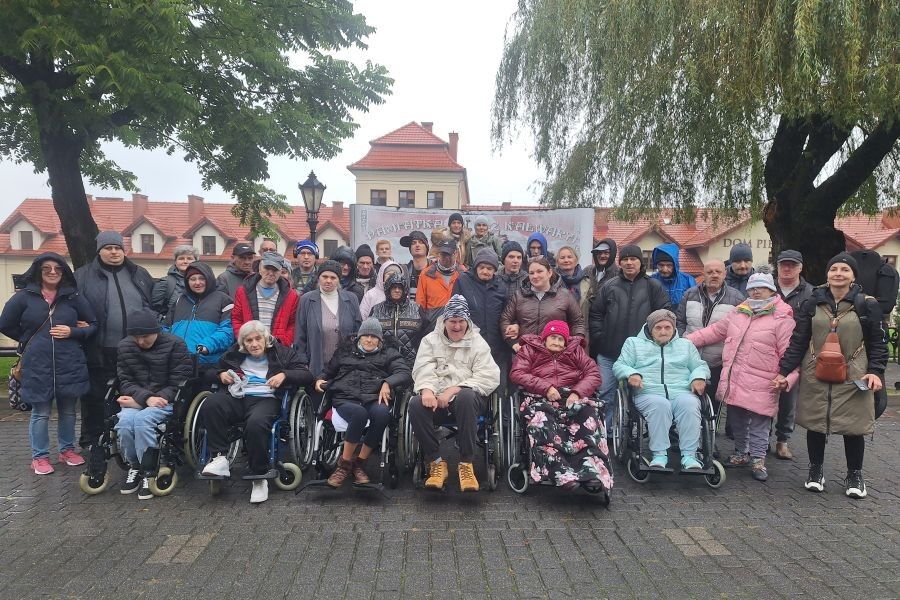 Grupa osób pozuje ustawiona w kilku rzędach do zdjęcia na tle budynków o czerwonych dachach. Zdjęcie wykonane na świeżym powietrzu w pochmurny dzień.