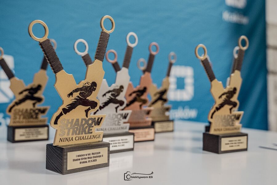 Zdjęcie przedstawia biały stół ze statuetkami dla zwycięzców zawodów Shadow Strike Ninja Challenge. Statuetki mają kształt skrzyżowanych kunai, a na ich złączeniu widać czarny rysunek ninja.