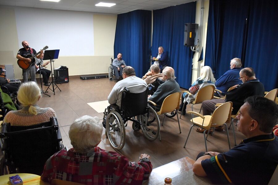 Grupa osób starszych, w tym kilka na wózkach inwalidzkich, siedzi w sali i słucha występu muzyka grającego na gitarze. Obok niego występuje drugi muzyk. 