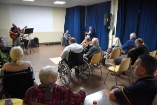 Grupa osób starszych, w tym kilka na wózkach inwalidzkich, siedzi w sali i słucha występu muzyka grającego na gitarze. Obok niego występuje drugi muzyk. 