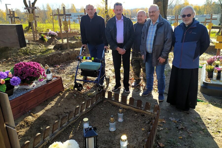 Pięciu mężczyzn stoi przy grobie na cmentarzu. Wokół widać inne groby, krzyże i kwiaty. Jeden z mężczyzn ma chodzik. Pogoda jest słoneczna, a drzewa w tle mają jesienne, żółte liście.