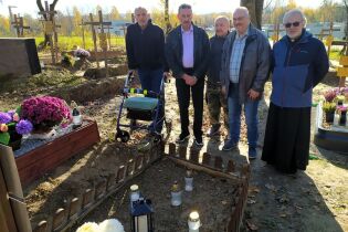 Pięciu mężczyzn stoi przy grobie na cmentarzu. Wokół widać inne groby, krzyże i kwiaty. Jeden z mężczyzn ma chodzik. Pogoda jest słoneczna, a drzewa w tle mają jesienne, żółte liście.