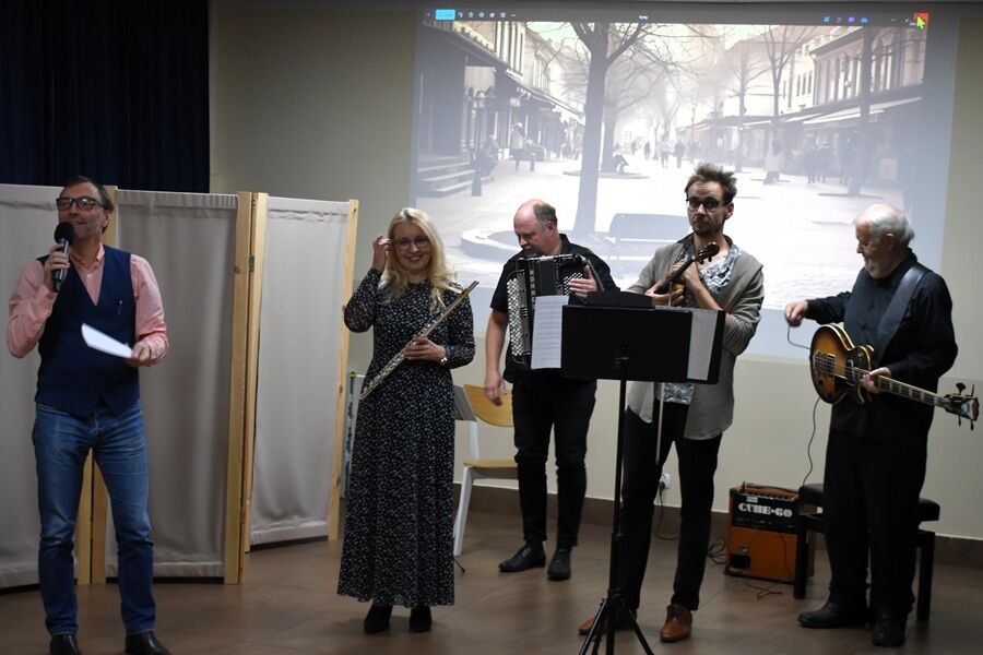  Grupa muzyków występuje na scenie w pomieszczeniu. Mężczyzna z mikrofonem mówi do publiczności, obok niego stoi kobieta z fletem oraz trzech muzyków grających na akordeonie, skrzypcach i gitarze.