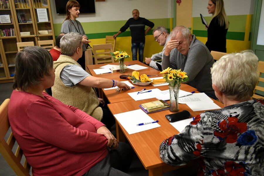 Kilka osób starszych siedzi przy stole, na którym leżą kartki papieru, długopisy i książki. Dwie kobiety stoją i prowadzą zajęcia. Na stole stoją wazony z żółtymi kwiatami.