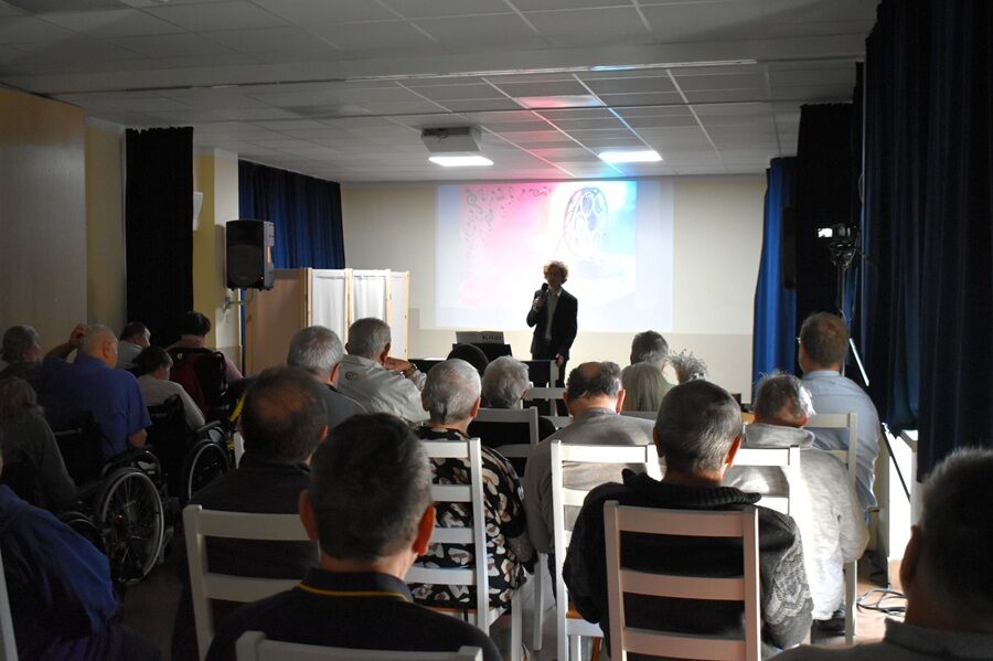 W sali z rzędem krzeseł i wózków inwalidzkich siedzi grupa starszych osób, oglądających występ mężczyzny z mikrofonem na tle ekranu z kolorową projekcją.