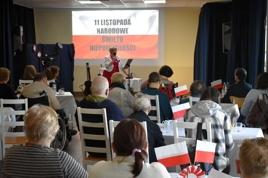 Sala ozdobiona polskimi flagami. Starsze osoby siedzą przy stołach, oglądając występ kobiety w białej marynarce i czerwonej sukience. Na ekranie w tle widać napis „11 LISTOPADA NARODOWE ŚWIĘTO NIEPODL