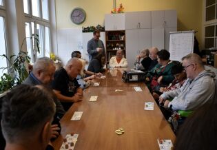 Grupa osób siedzi wokół dużego stołu w jasnym pomieszczeniu, grając w grę planszową lub lotto. Na ścianie widać zegar i szafki z kolorowymi dekoracjami, a w tle stoi mężczyzna prowadzący zajęcia.