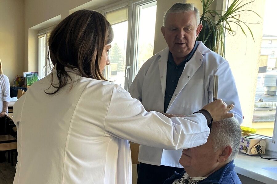 Kobieta i mężczyzna w białych fartuchach wykonują badanie lub pomiar głowy starszego mężczyzny w jasnym pokoju z dużymi oknami. W tle widać inne osoby i wyposażenie gabinetu.