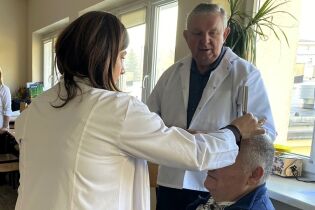 Kobieta i mężczyzna w białych fartuchach wykonują badanie lub pomiar głowy starszego mężczyzny w jasnym pokoju z dużymi oknami. W tle widać inne osoby i wyposażenie gabinetu.