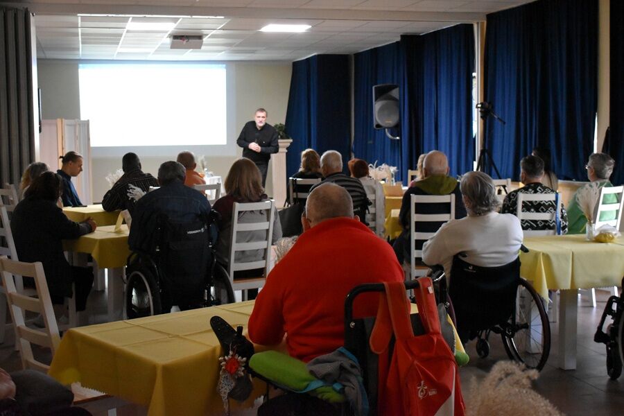 Sala z osobami siedzącymi przy stołach przykrytych żółtymi obrusami, część uczestników na wózkach inwalidzkich. Na scenie przed ekranem stoi mężczyzna prowadzący prezentację lub wykład.
