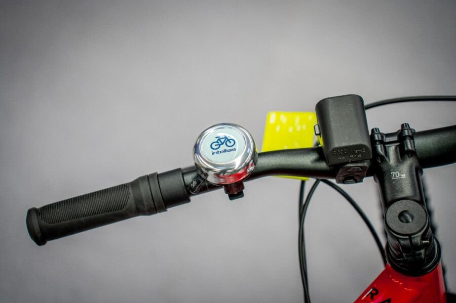 Fragment czerwonego roweru z czarną kierownicą. Na kierownicy znajduje się dzwonek i przednia lampka, typowe akcesoria roweru.